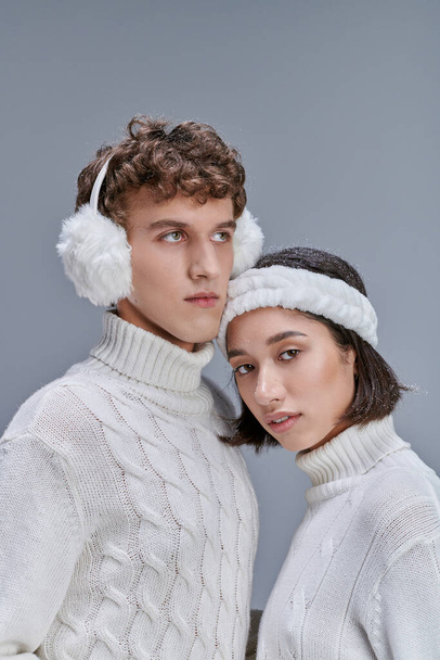 молода азіатка зі сніжним волоссям дивиться на камеру біля чоловіка в теплих вухах на сірому, зимовому стилі - Фото, зображення