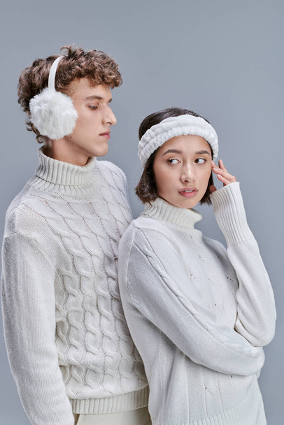 stylish asian woman looking away near young man in warm earmuffs on grey background, snowy hair - Photo, Image