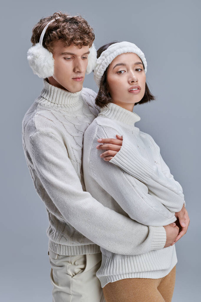 trendy man in earmuffs hugging young asian woman on grey background, stylish romantic winter - Photo, Image