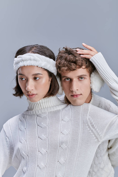 aziatische vrouw aanraken besneeuwde haar van de man in warme oorkappen op grijze achtergrond, romantische winter stijl - Foto, afbeelding