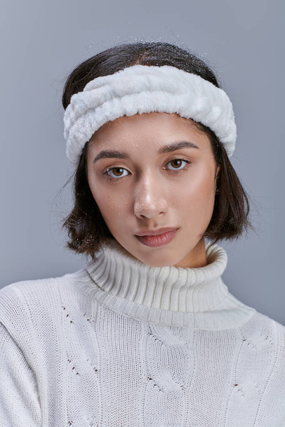 portrét mladé asijské ženy v teplé čelence a bílý pletený svetr při pohledu na kameru na šedé - Fotografie, Obrázek