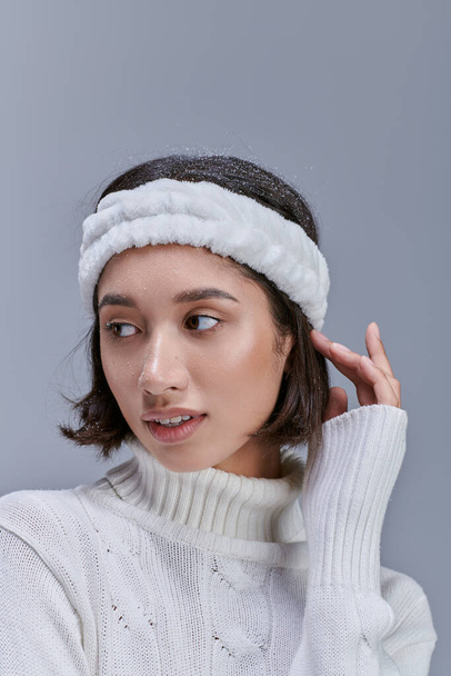 stylish asian woman in warm headband and white sweater posing on grey, winter fashion concept - Photo, Image