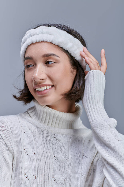 téli divat kampány, vidám ázsiai modell kötött pulóverben és fejpántban, félrenézve a szürkére - Fotó, kép