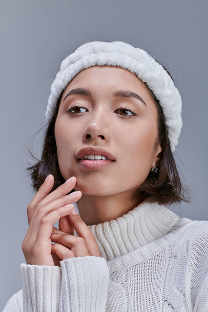 romantisch en dromerig aziatisch vrouw in de winter trui en warm wit hoofdband weg te kijken op grijs - Foto, afbeelding
