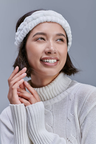 portrait of carefree asian woman in knitted sweater headband looking away on grey, happy winter - Photo, Image