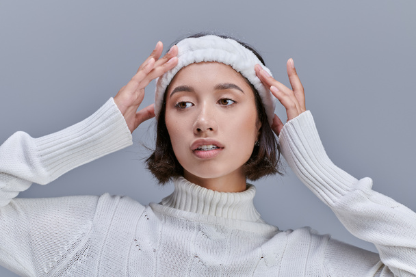 stylish asian woman in knitted sweater adjusting warm headband on grey, winter fashion photography - Photo, Image