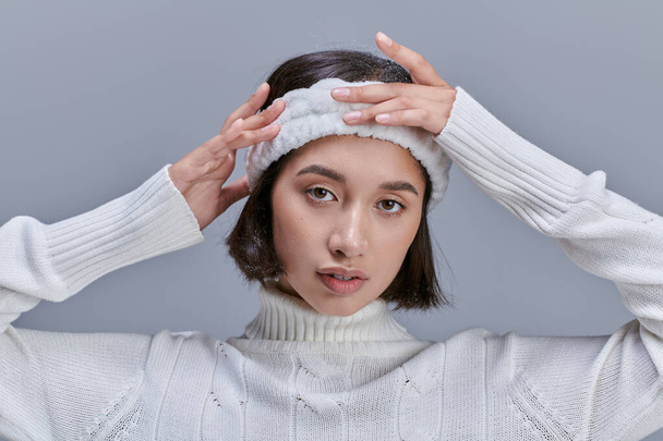 jong aziatische vrouw in wit trui aanpassen zachte hoofdband op grijs, winter mode concept - Foto, afbeelding