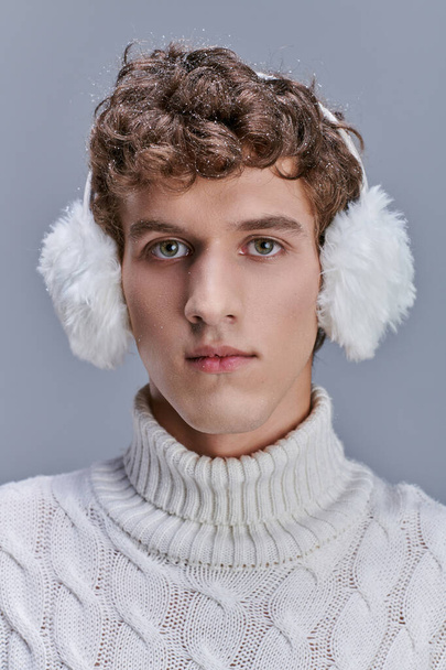 ritratto di giovane uomo con neve sui capelli ondulati in posa in caldo paraorecchie e maglione bianco su grigio - Foto, immagini