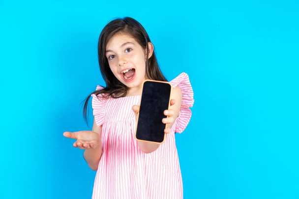 kid with a mobile phone. presenting smartphone. Advertisement concept. Beautiful caucasian little girl posing over blue studio background - Photo, Image