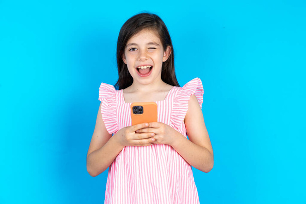 Excited kid winking and eye hold smart phone use read social network news. Beautiful caucasian little girl posing over blue studio background - Photo, Image