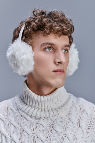 homme élégant avec de la neige sur les cheveux ondulés, en cache-oreilles et pull blanc regardant loin sur gris, portrait - Photo, image
