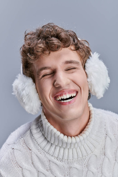 opgewonden man in oorkappen en witte trui lachen met gesloten ogen op grijs, gelukkig stijlvolle winter - Foto, afbeelding
