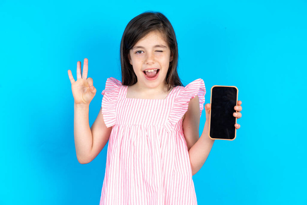 Το παιδί κρατάει το κινητό του και δείχνει εντάξει. Όμορφο καυκάσιο κοριτσάκι που ποζάρει πάνω από το μπλε φόντο του στούντιο - Φωτογραφία, εικόνα