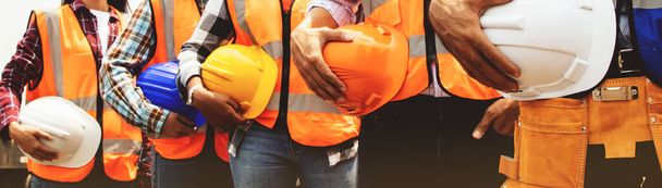 Inżynierowie całego zespołu mężczyźni i kobiety pracownicy różnych narodowości noszący kamizelki stojące w kolejce trzymające hełmy bezpieczeństwa w celu zapewnienia bezpieczeństwa i zapobiegania wypadkom podczas pracy w przemyśle fabrycznym. - Zdjęcie, obraz