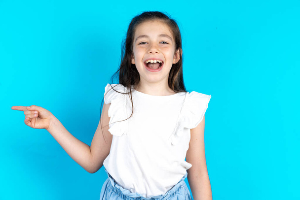 Ребенок смеется счастливо указывает прочь на пустое пространство демонстрирует торговые скидки предложение, взволнованный хорошей новостью или неожиданной продажи. Красивая кавказская маленькая девочка позирует на фоне голубой студии - Фото, изображение