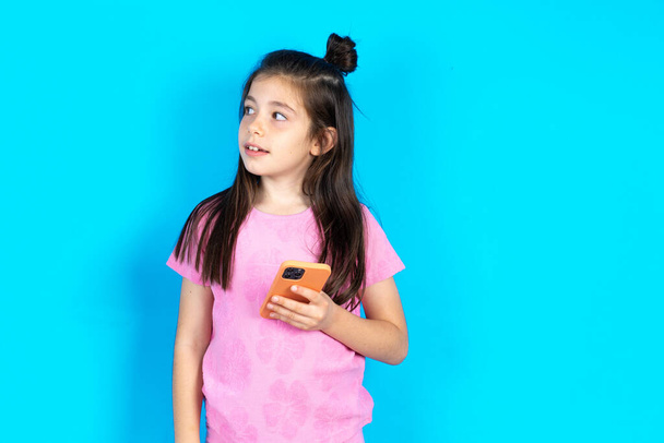 Kind besitzt ein Mobiltelefon, nutzt schnelles Internet und soziale Netzwerke, Konzept der Online-Kommunikation und moderne Technologien. Schöne kaukasische kleines Mädchen posiert über blauem Studiohintergrund - Foto, Bild