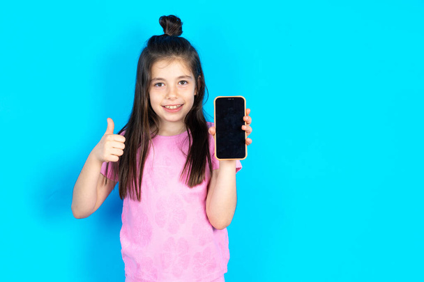 Kid Showing blank screen smartphone, thumb up, recommending new app. Beautiful caucasian little girl posing over blue studio background - Photo, Image