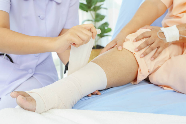 Lähikuva naispuolisen sairaanhoitajan käsi hoitaa iäkästä naispotilasta, joka on sairaalahoidossa nilkkakivun vuoksi nyrjähtänyt jalka sairaanhoitajat käyttävät siteitä lievittää jalka ja nilkka kipua potilaan sängyssä: sairausvakuutus. - Valokuva, kuva