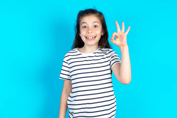 szczęśliwy uśmiechnięty dzieciak pokazuje okey symbol z jej ręki. piękne biały dziewczyna pozowanie nad niebieski studio tło  - Zdjęcie, obraz