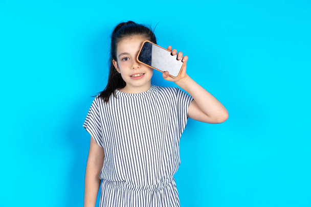Dzieciak trzymający nowoczesny smartfon, zakrywający jedno oko przy uśmiechu. Piękny biały dziewczynka w paski t-shirt stoi nad niebieskim tle studio  - Zdjęcie, obraz