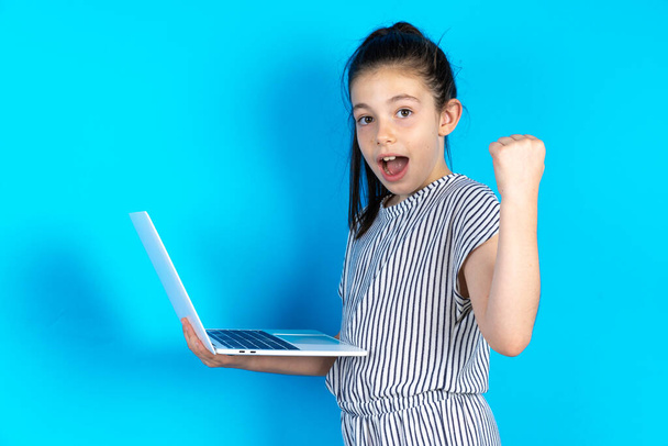 Красивая белая маленькая девочка в полосатой футболке стоит на синем фоне студии, держа в руках компьютер, открывая рот, поднимая кулак - Фото, изображение
