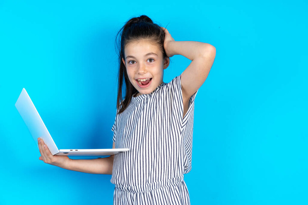 Фото изумленной белой маленькой девочки в полосатой футболке, стоящей на синем фоне студии с современным гаджетом, рука об голову - Фото, изображение