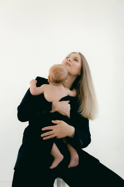 Jonge vrouw met baby in haar armen. Blond meisje in zwarte kleren glimlachend naar uw kind op een witte achtergrond. Gelukkig moederschap en borstvoeding concept. Foto 's met zonneschijn, zachte focus, overbelichting. - Foto, afbeelding