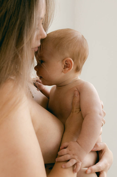 Femme nue avec bébé dans les bras sur fond clair. Joyeux concept de maternité et d'allaitement. Photos avec mise au point douce. Bonjour, concept. Bébé nu mignon regardant la caméra. - Photo, image