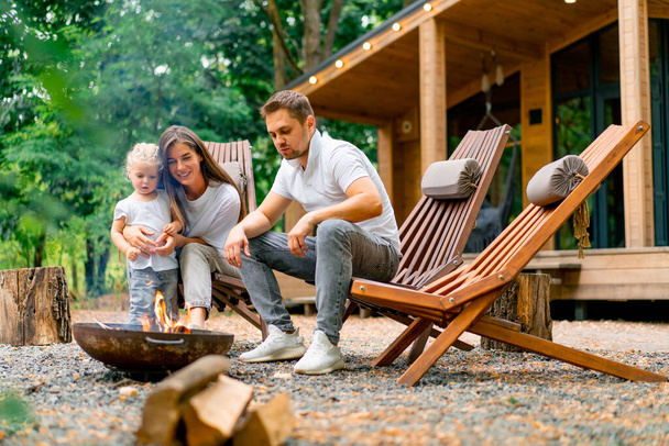 Bonito jovem família pai mãe e filha estão sentados juntos por um incêndio na floresta e assar marshmallows perto de sua casa de campo - Foto, Imagem