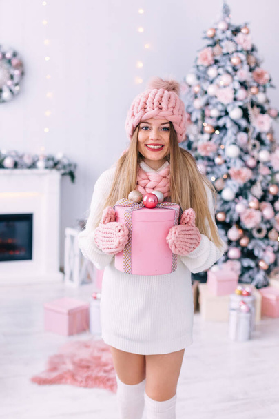 Örgü atkı, şapka ve eldiven takmış şık bir kız bir hediye tutuyor ve kameraya bakıyor. Şömineli ve Noel ağacı olan oda.. - Fotoğraf, Görsel