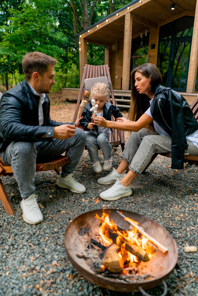 Cute young family dad mom and daughter are sitting together by a fire in the forest and roasting marshmallows near their country house - Photo, Image