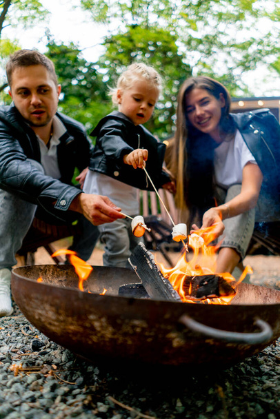 Primer plano de un padre de familia joven, mamá e hija sentadas juntas junto a un fuego en el bosque y asando malvaviscos cerca de su casa de campo - Foto, Imagen