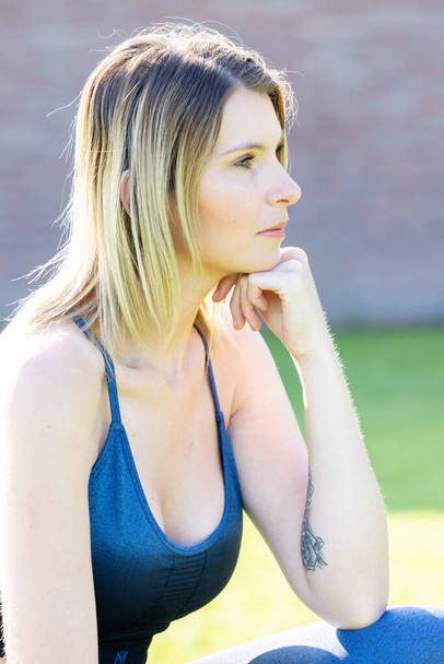 Une jeune femme millénaire séduisante et parfaitement formée en tenue de sport bleue prend un moment pour se reposer sur l'herbe à l'extérieur, en regardant doucement loin de la caméra. Sporty Millennial Femme reposant sur - Photo, image