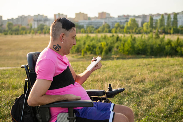 niepełnosprawny gej człowiek siedzi w a wózek inwalidzki i używa a telefon komórkowy działa online. - Zdjęcie, obraz