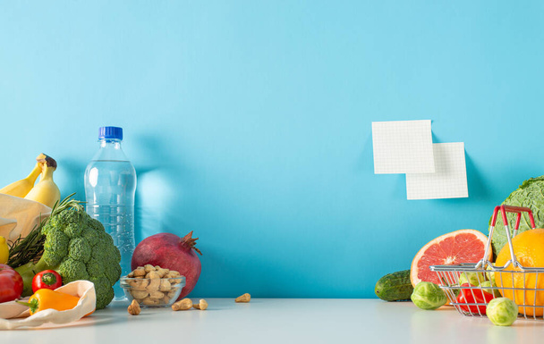 Ρύθμιση της αγοράς με επίκεντρο την υγεία: πίνακας πλευρικής προβολής με νερό, ξηρούς καρπούς, λαχανικά, φρούτα όπως μπανάνες, λάχανο, μπρόκολο και γκρέιπφρουτ. Μπλε τοίχο φόντο με κολλώδη χαρτιά σημείωμα και χώρο για διαφημίσεις - Φωτογραφία, εικόνα