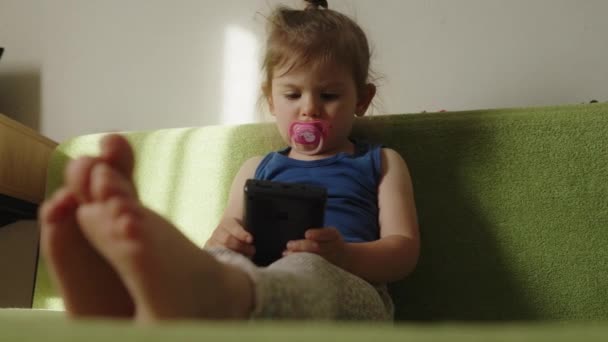 Малышка сосет соску, играя в портативную видеоигру, сидя на зеленом диване в гостиной. Выходные дни - Кадры, видео