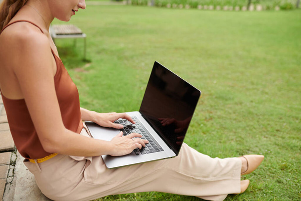 Female freelancer sitting on lawn in park and working on laptop, answering e-mails from clients - Photo, Image