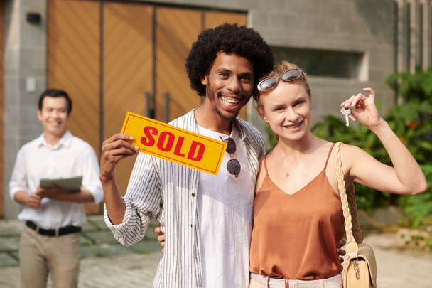 Portrait of happy excited diverse couple showing sold sign and keys from new house - Photo, Image