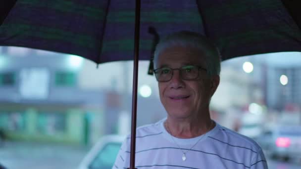 Een oudere man wandelen in Rainy City Sidewalk met paraplu glimlachend - Video