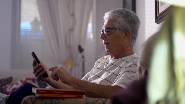 Un anciano feliz usando un celular en casa sentado en el sofá. Cabello gris hombre mayor caucásico persona comprometida con la tecnología moderna, navegar por Internet en línea - Imágenes, Vídeo
