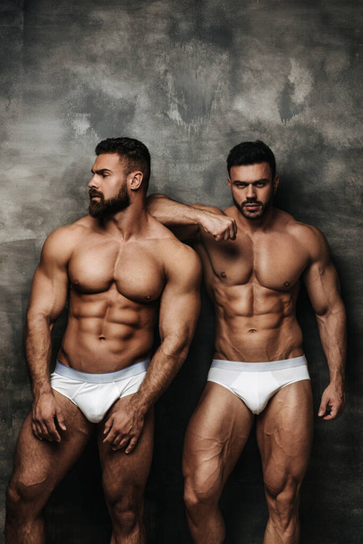 Двоє м'язистих чоловіків в білій білизні і позує в студії. Двоє сексуальних бодібілдерів стоять біля стіни. Пара красивих фітнес-хлопців. - Фото, зображення