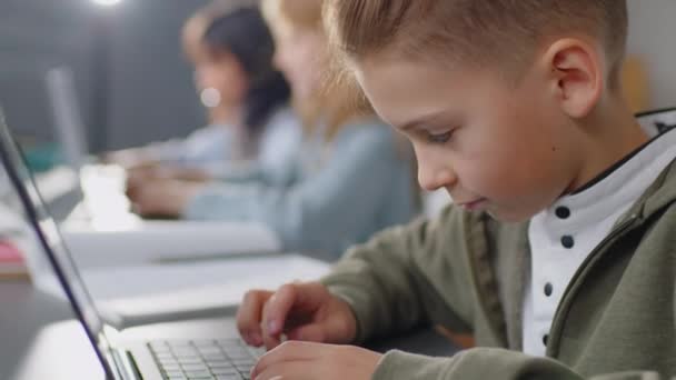 Enfoque inclinado en el niño de edad elemental caucásico sentado en el escritorio en el aula escribiendo con atención algo en el portátil durante el día - Imágenes, Vídeo