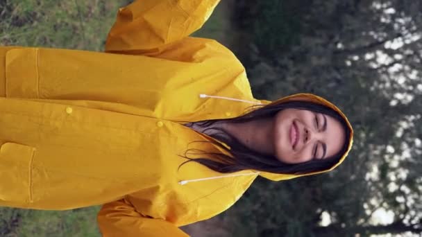 retrato de una joven sonriente con un impermeable amarillo durante la lluvia en el parque. Mujer alegre disfrutando de la lluvia al aire libre y bailando. expresión alegre en su rostro mientras disfruta del clima. vídeo vertical - Imágenes, Vídeo