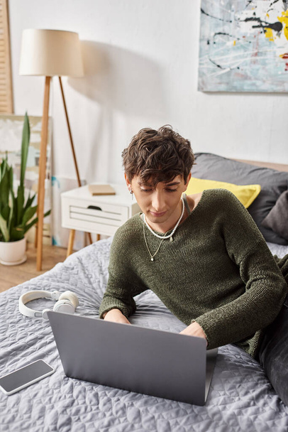 young curly transgender blogger using laptop and sitting on bed near smartphone and headphones - Photo, Image