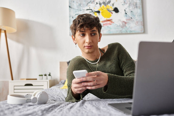 junge lockige Transgender-Bloggerin mit Smartphone in der Nähe von Laptop und auf dem Bett in der Nähe von Kopfhörern - Foto, Bild
