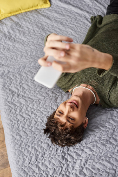 ευτυχισμένος τρανσέξουαλ blogger με σγουρά μαλλιά χρησιμοποιώντας smartphone και ξαπλωμένος στο κρεβάτι, κοινωνική επιρροή των μέσων μαζικής ενημέρωσης - Φωτογραφία, εικόνα