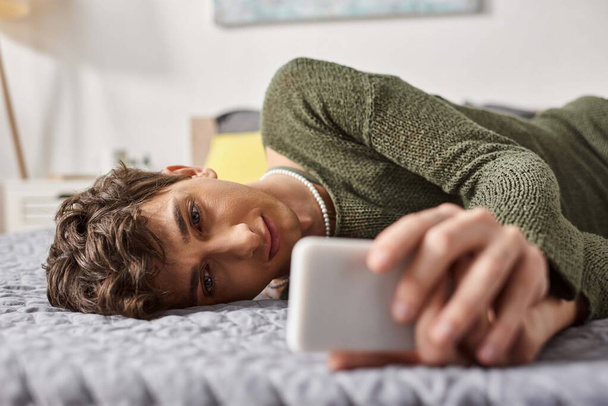 Τρανσέξουαλ blogger με σγουρά μαλλιά χρησιμοποιώντας smartphone και ξαπλωμένος στο κρεβάτι, επιρροή στα μέσα κοινωνικής δικτύωσης - Φωτογραφία, εικόνα