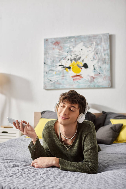 šťastný kudrnatý transsexuál v bezdrátových sluchátkách držící smartphone v ložnici, poslouchat hudbu - Fotografie, Obrázek