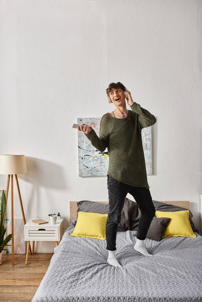 feliz persona transexual rizada en auriculares inalámbricos sosteniendo el teléfono inteligente y de pie en la cama - Foto, Imagen
