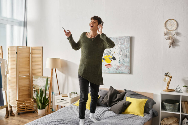 szczęśliwy kręcone transseksualista osoba w bezprzewodowy słuchawki trzymając smartfon i stojąc na łóżku - Zdjęcie, obraz